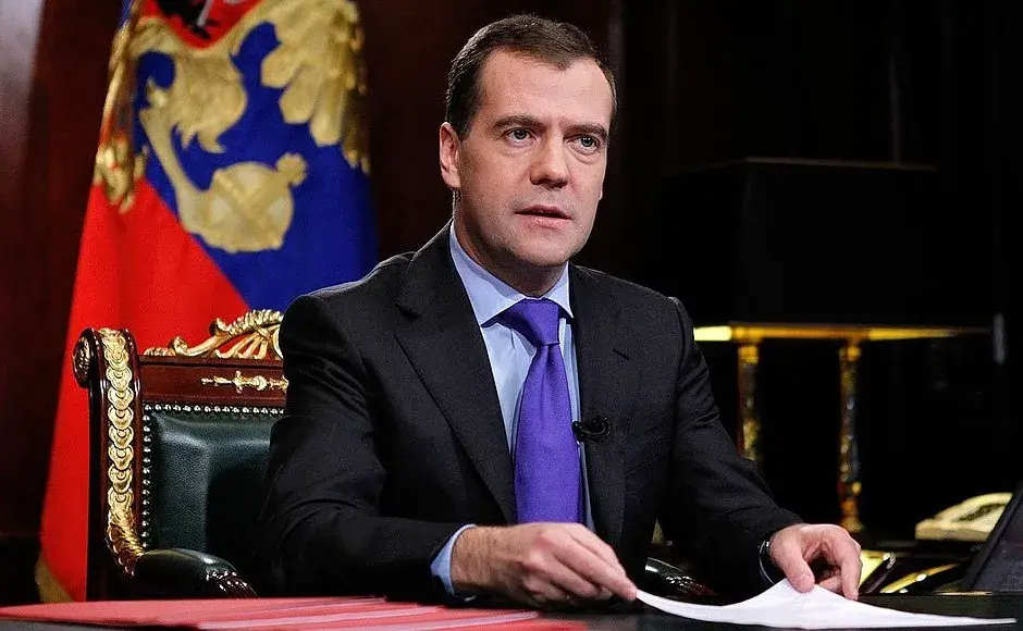 Dmitry Medvedev sitting with a Russian flag behind him.