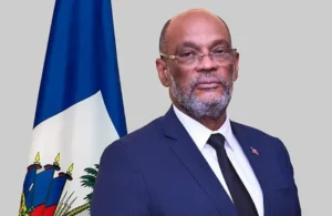 Former Prime Minister of Haiti, Ariel Henry, standing with the flag of Haiti behind him.